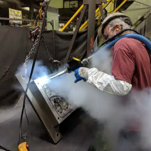 Operador com equipamento de proteção a limpar um molde metálico com jato de gelo seco, com nuvem de CO₂ visível